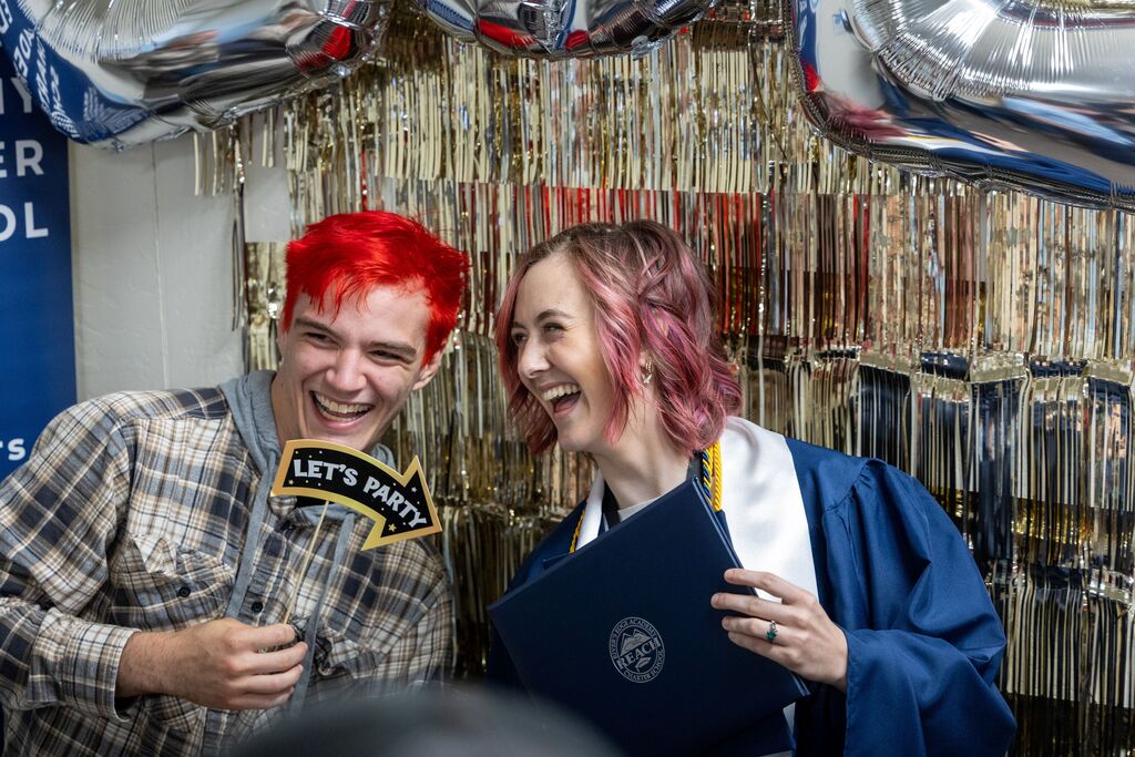 A graduating senior young woman holds her diploma in front of a sparkling photo wall. She laughs. Next to her, a young man holds a "Let's Party!" sign. He also laughs. 