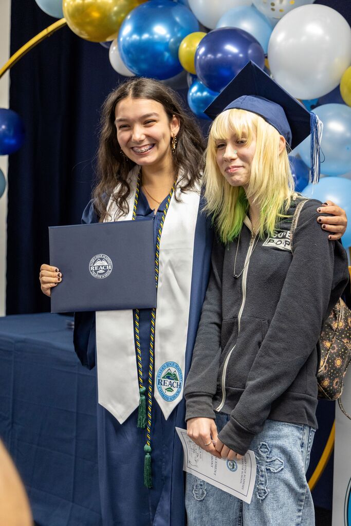 A graduating senior young women holds her diploma for a picture. Next to her, a teenage girl wears the graduate's cap. 