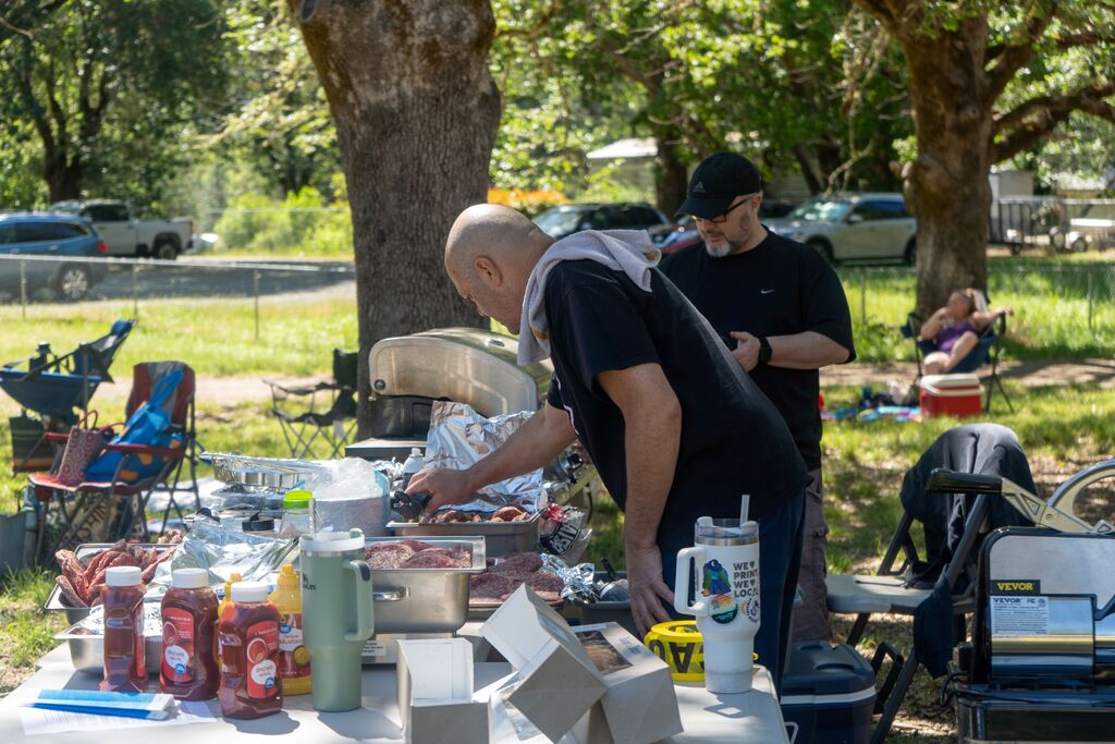 Two men  put raw hamburgers onto a grill. 