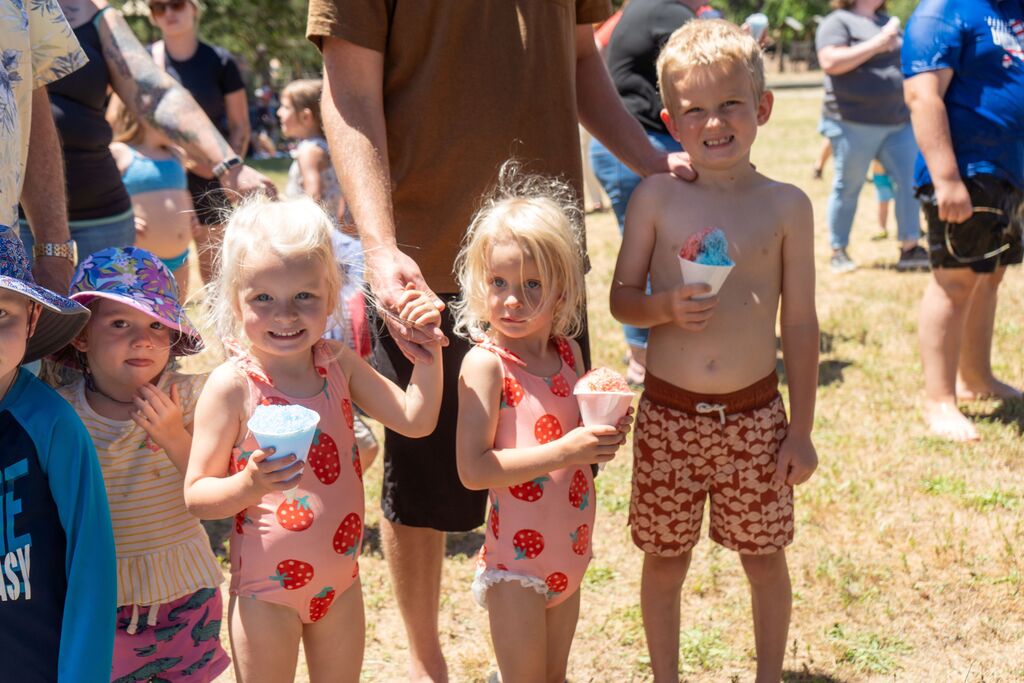 3 small children smile at the camera. They are wearing swimsuits and holding snowcones. 