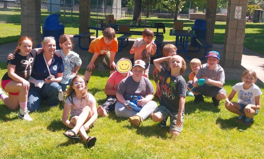 Several children kneel on grass and smile at the camera. A teacher kneels alongside them. 