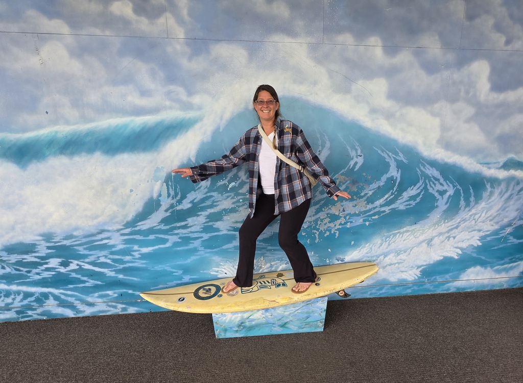 A woman poses on a surfboard in front of a wave painted on a wall. 