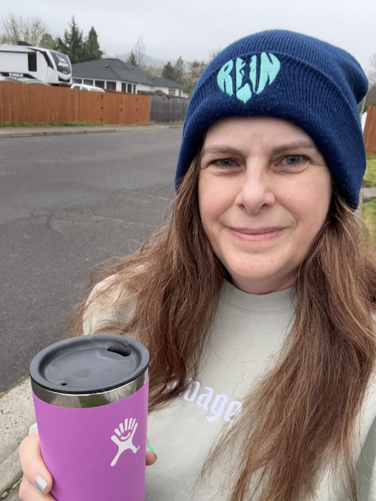 A smiling woman wears a beanie and holds a purple drink tumbler. 