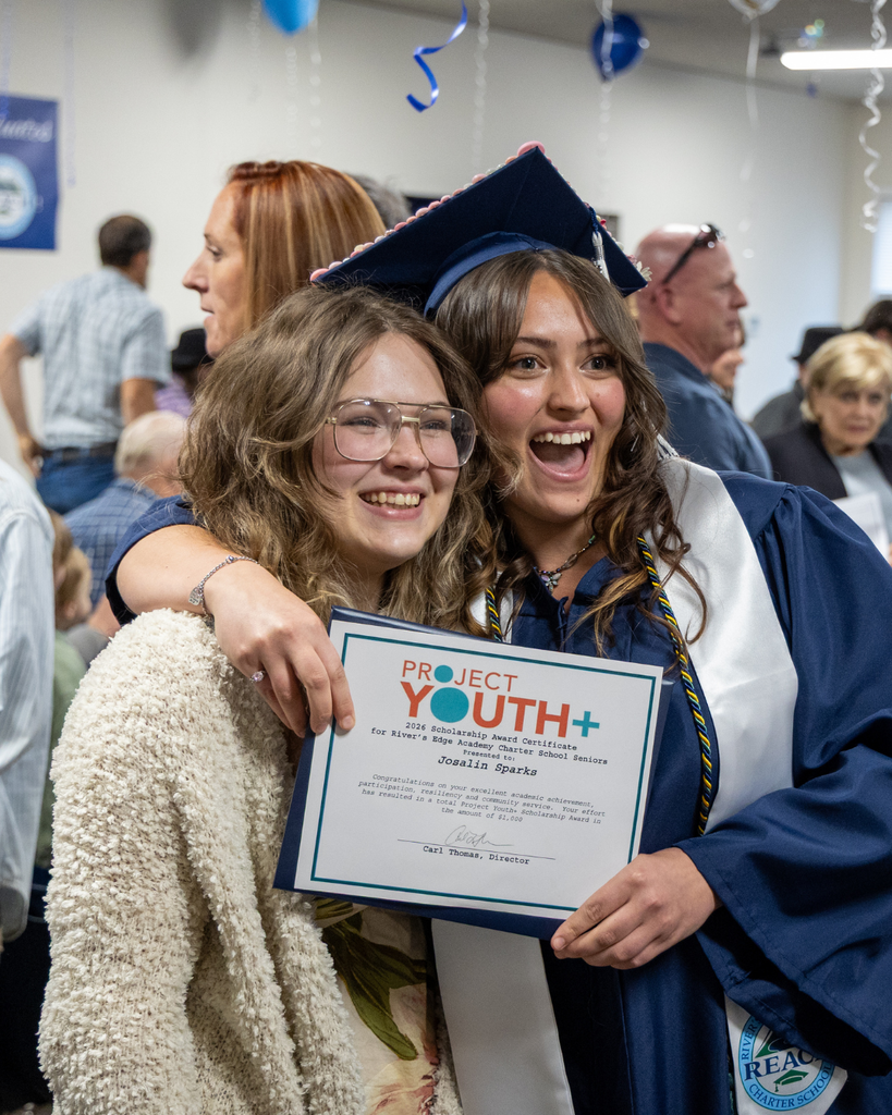 A young woman in graduation attire holds a certificate with her arm wrapped around another young woman. They are smiling at a camera.
