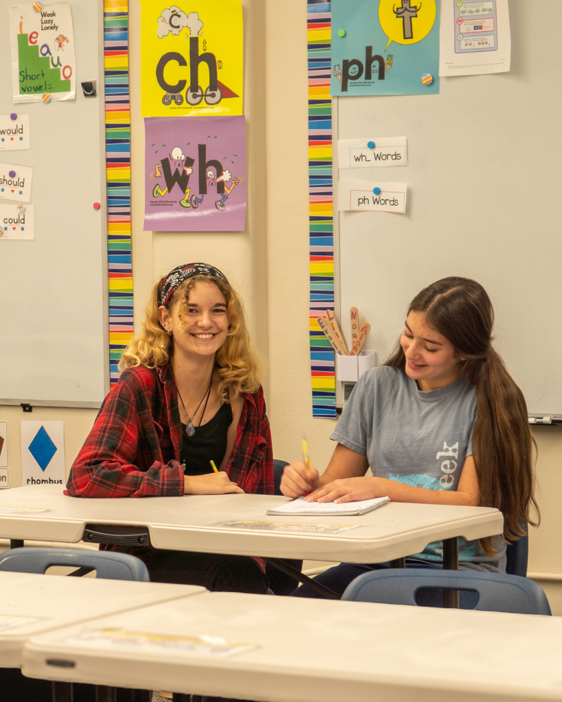 Two smiling high school girls sit a desk. They are writing on a piece of paper. 