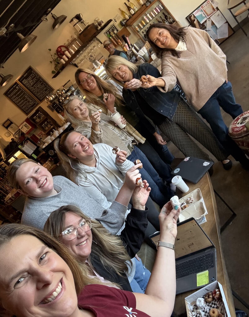 Seven ladies hold donuts and stand around a table, smiling while taking a selfie. 