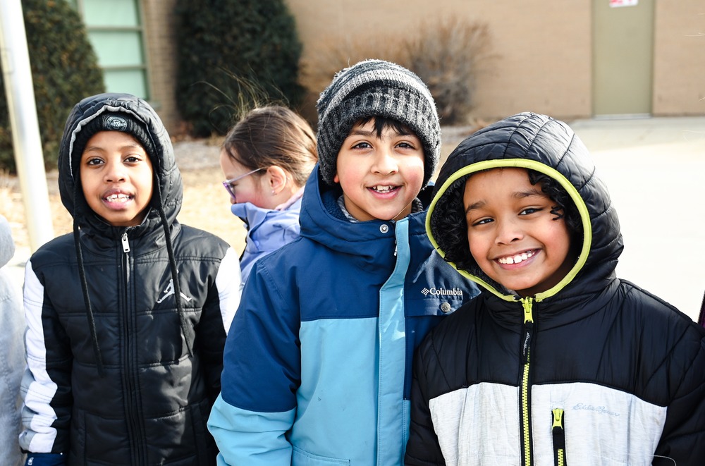 Elementary schoolers in their winter coats and hats smile at the camera.