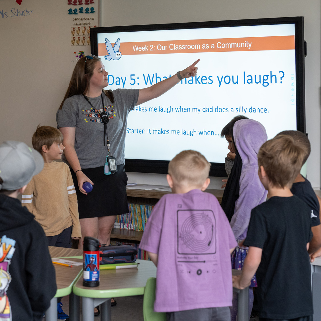Teacher points to a smartboard that says, "Week 2: Our Classroom as a Community; Day 5: What makes you laugh?"
