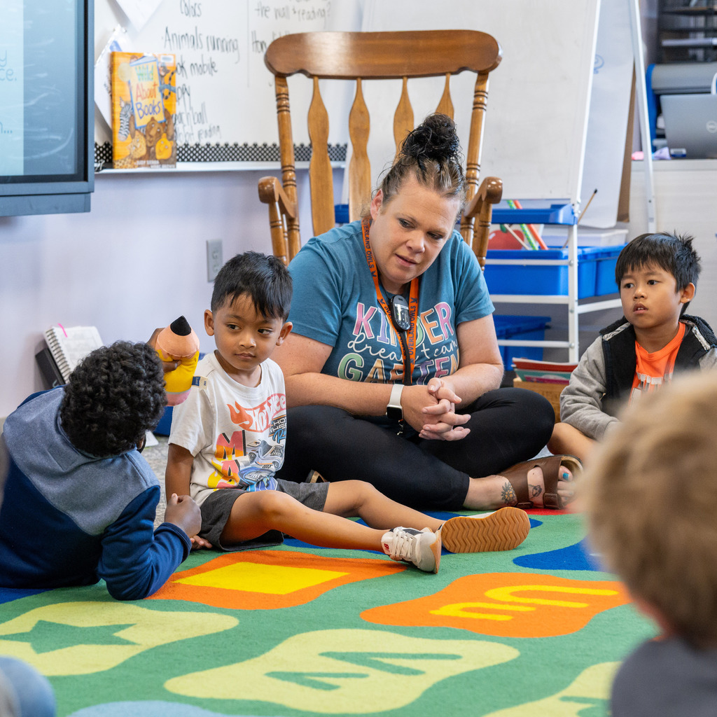 Teacher and students sit on the floor and listen to the kid with the plush pencil talk.