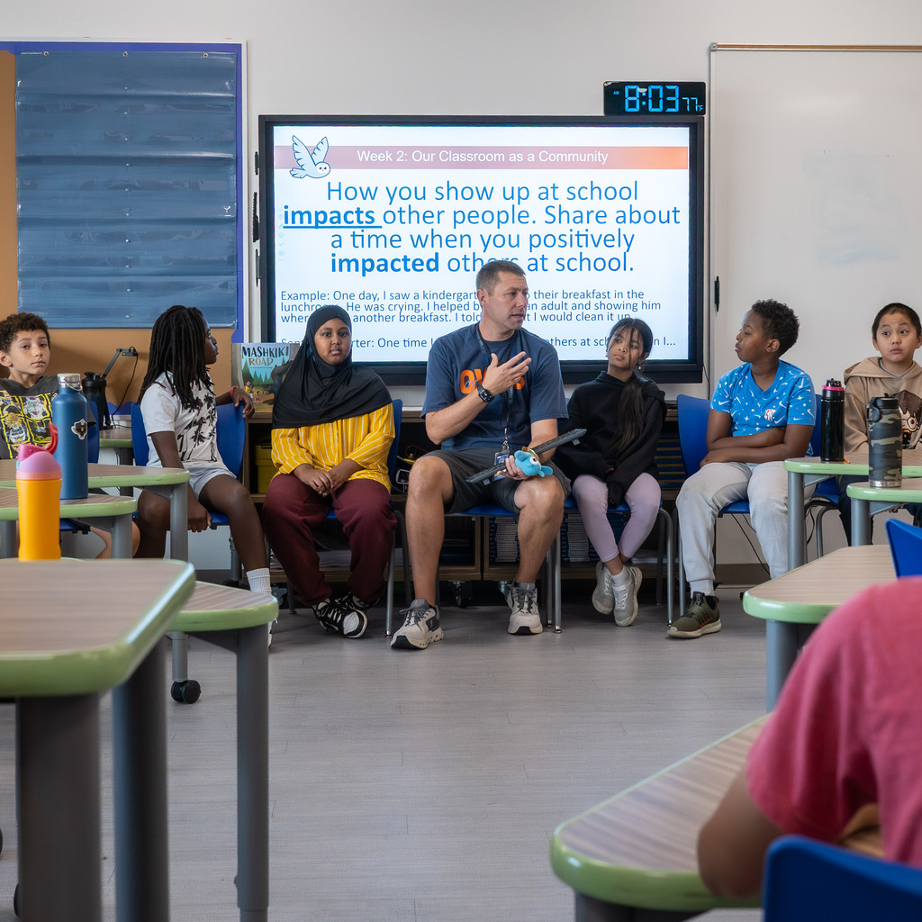 Teacher and class sit in a  discussion circle with a prompt on the smartboard that says, "How you show  up at school impacts other people. Share about a time when you positively impacted others at school."