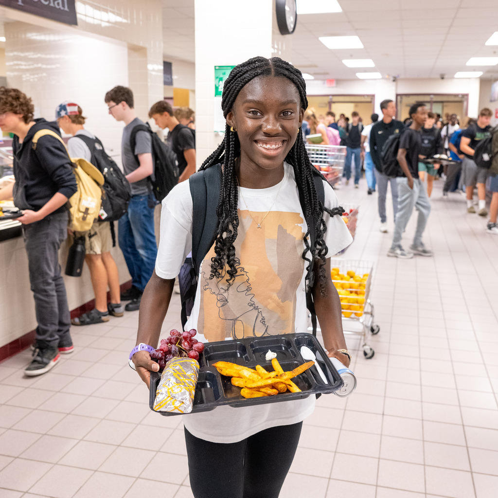 High school student in cafeteria smiling and holding her lunch tray filled with fries, grapes, and a wrap.