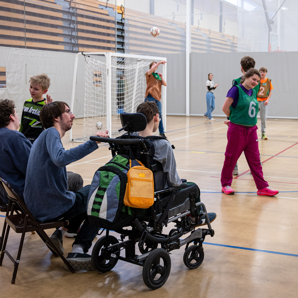 Two onlookers sit in chairs and one in a wheelchair as they watch a unified soccer game.
