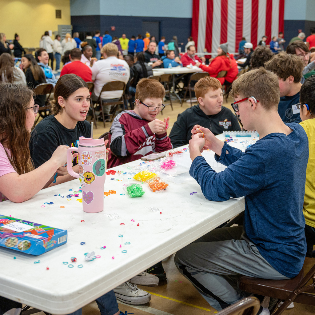 Students loop small rubber bands together as a craft at a fold-out table.