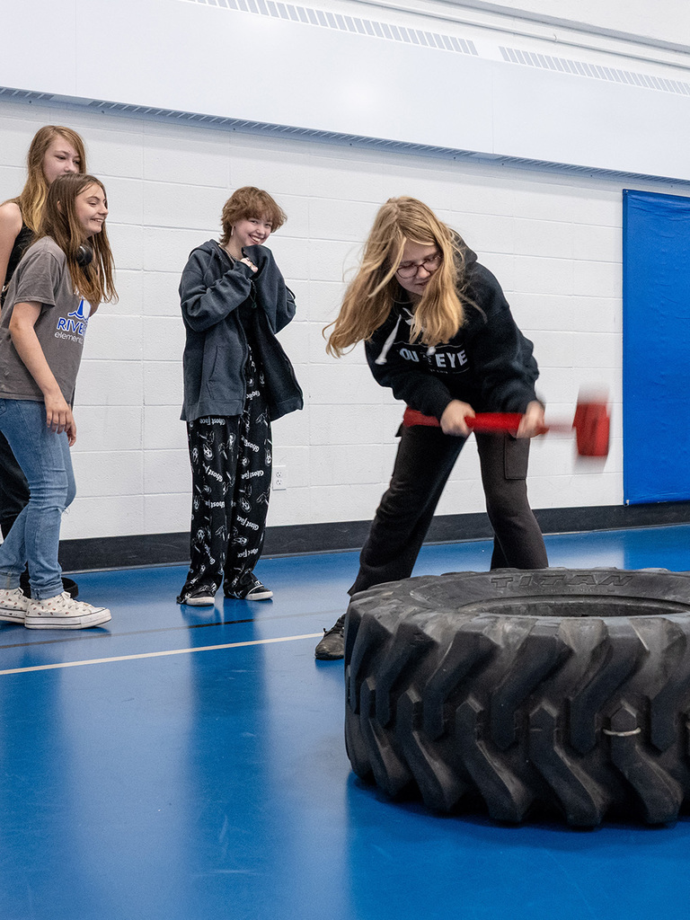 Girl's friends look on smiling while she whacks a tire with a mallet.