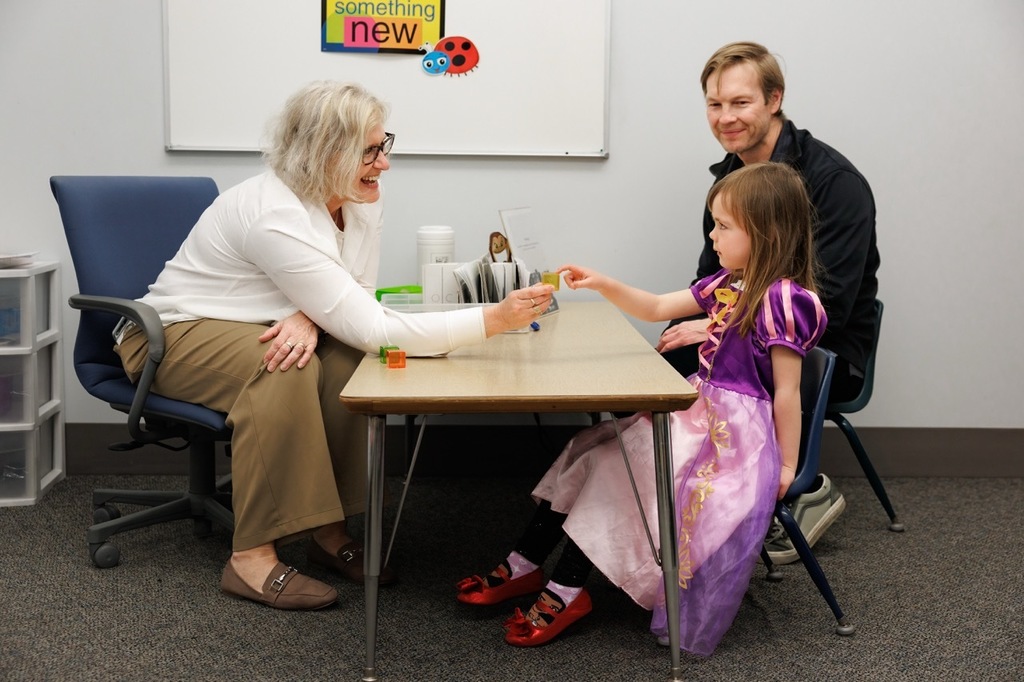 Early Childhood screener holding a block toward a child smiling while the dad is in the background smiling too.