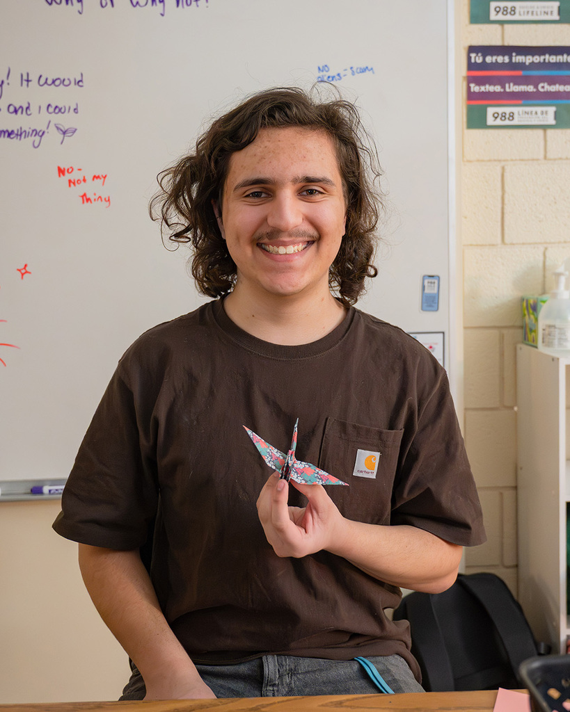 A high school student smiles at the camera holding a colorful paper crane he just folded.