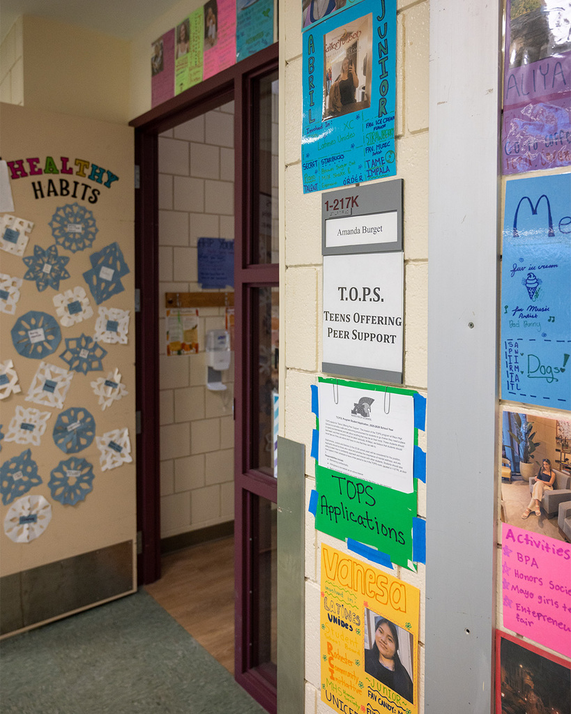 Outside an open door are hung decorative handmade bios of Peer Supporters on colorful construction paper. The sign by the door reads, "T.O.P.S. Teens Offering Peer Support."