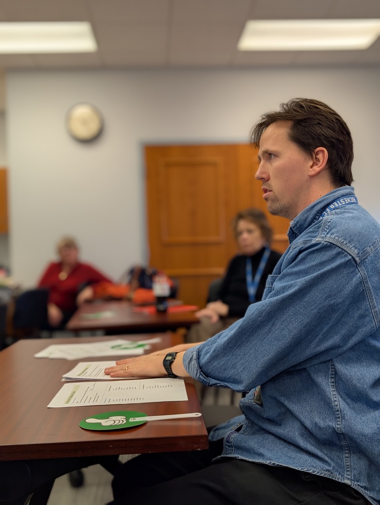 A man in a denim shirt is transfixed by the speaker. Handouts and a thumbs-up paddle sit on the desk in front of him.