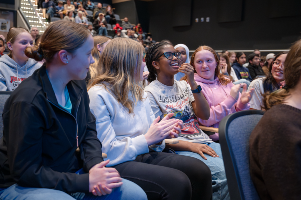 Student smiling after hearing they had won, surrounded by peers clapping and smiling at her. 