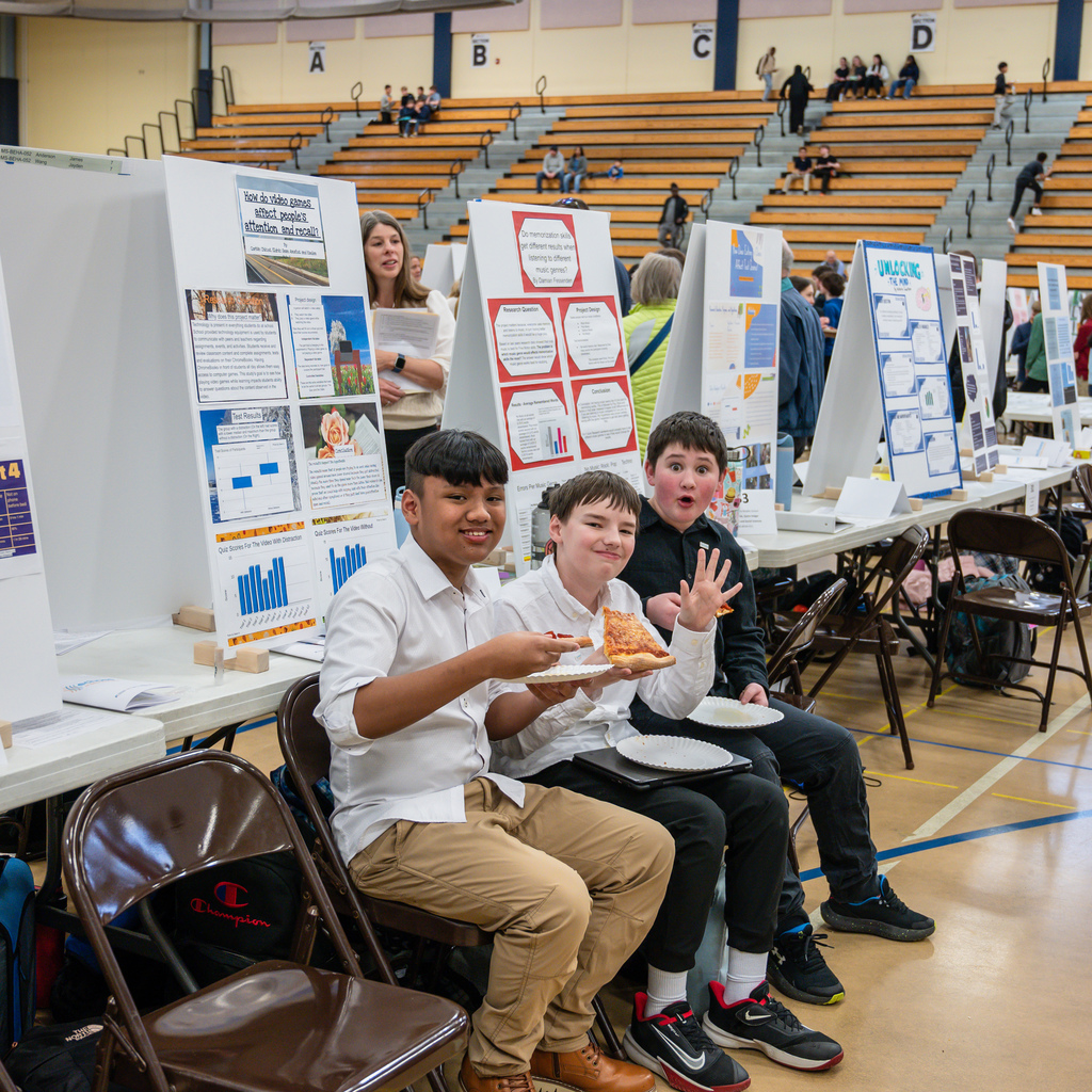 Three STEM fair participants taking a lunch break while holding pizza in their hand and waving to the camera.