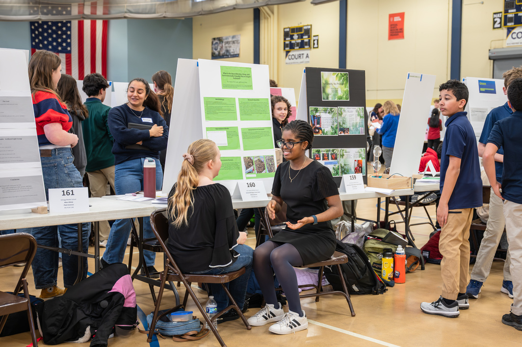 Student talking to Judge while sitting in chairs infront of the project board. 