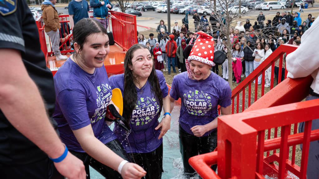 Three high school girls wearing smiles and matching "Polar Plunge" t-shirts emerge from the frigid waters of the Polar Plunge tank. They are flanked by a life guard and a police officer.