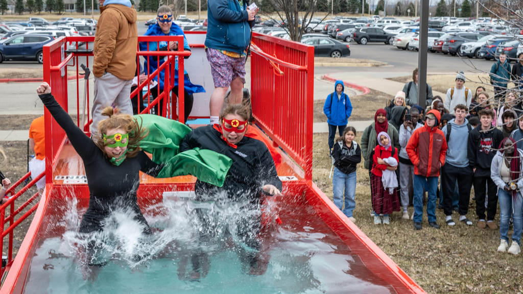 Two RPS staff members in capes and masks jump into the Polar Plunge. In the background, students look on from the ground close to a parking lot full of cars.