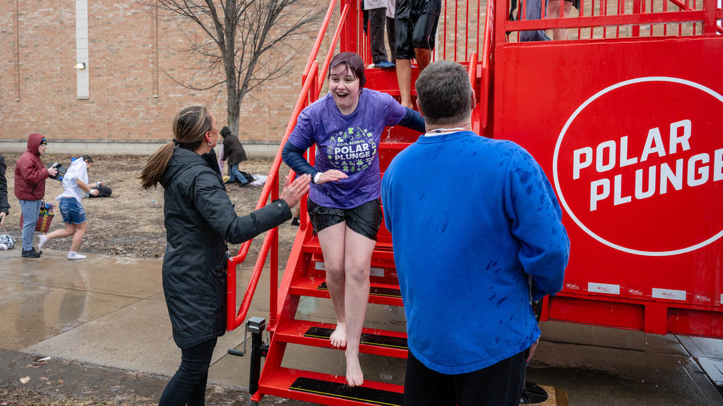Two adults help a soaked student down from the Polar Plunge tank. She is cold but smiling through it.