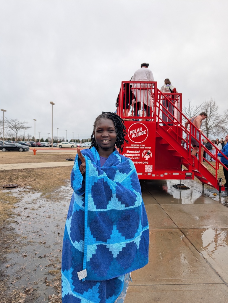 A student wrapped in a towel holds up a peace sign for the camera. The bright red Polar Plunge tank is in the background.