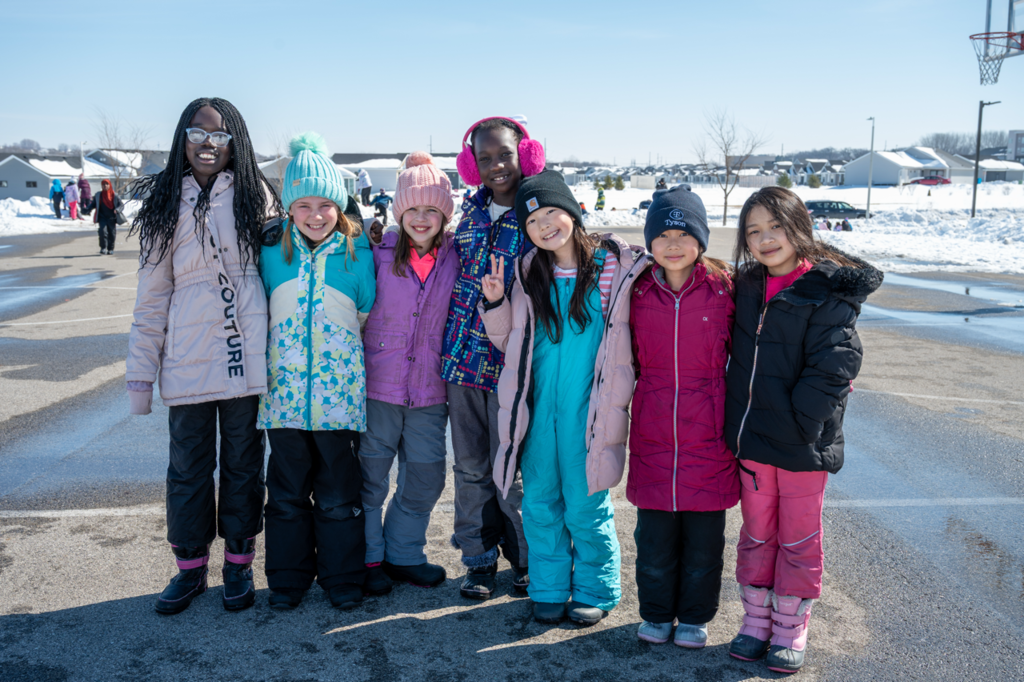 Group of seven students decked out in snow gear arm in arm on the black top for a photo
