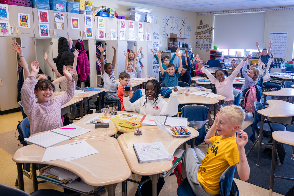 All the class siting at their desks with their hands in the air celebrating the photo and smiling at the camera.