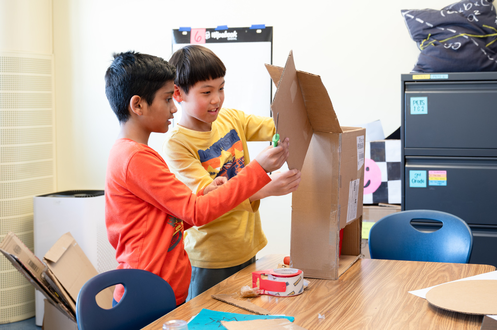 Two students building carnival games out of cardboard at their desks.