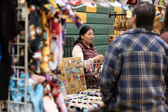 A Community vendor at our American Indian Education powwow set up behind a table filled with handmade clothing, beads, bracelets, and purses, while customers look at her products.