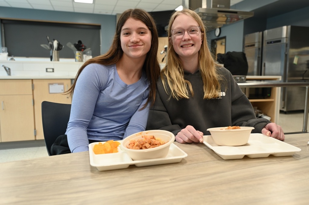 Two students sitting together with their food at the table smiling at the camera.