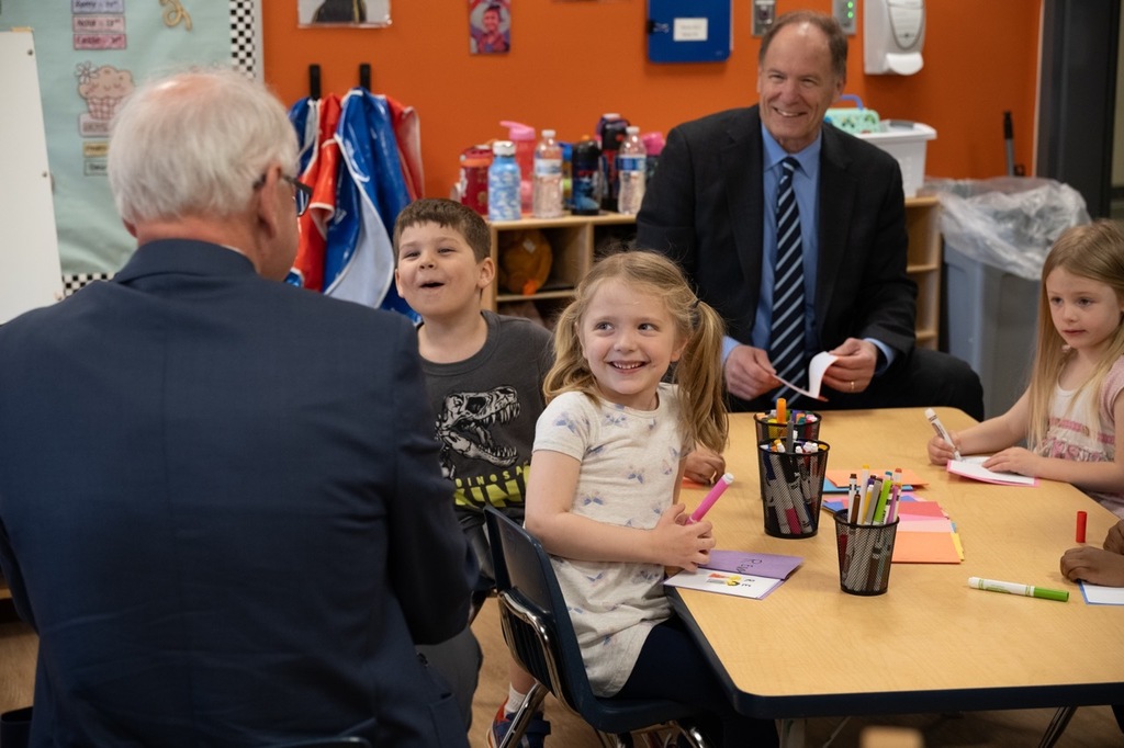 Governor Walz talking with a group of preschool students making story books about dinosaurs while the students look at Walz and smile or laugh with Commissioner Marquart smiling at the other end of the table.