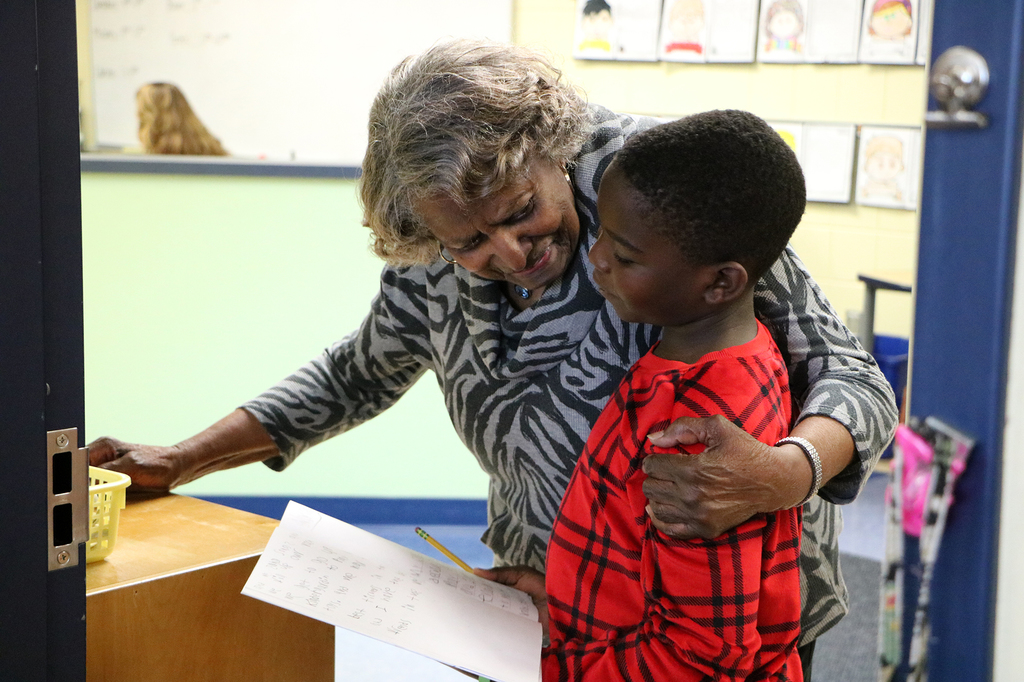 Mrs. Joyce Gibbs encouraging a student with her arm around them as they show her their work. 