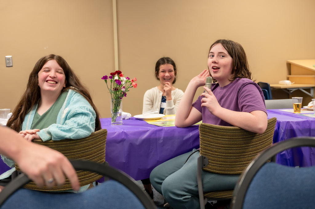 A student holding a microphone shares about their experience at camp while their peer across the table laughs at the story.