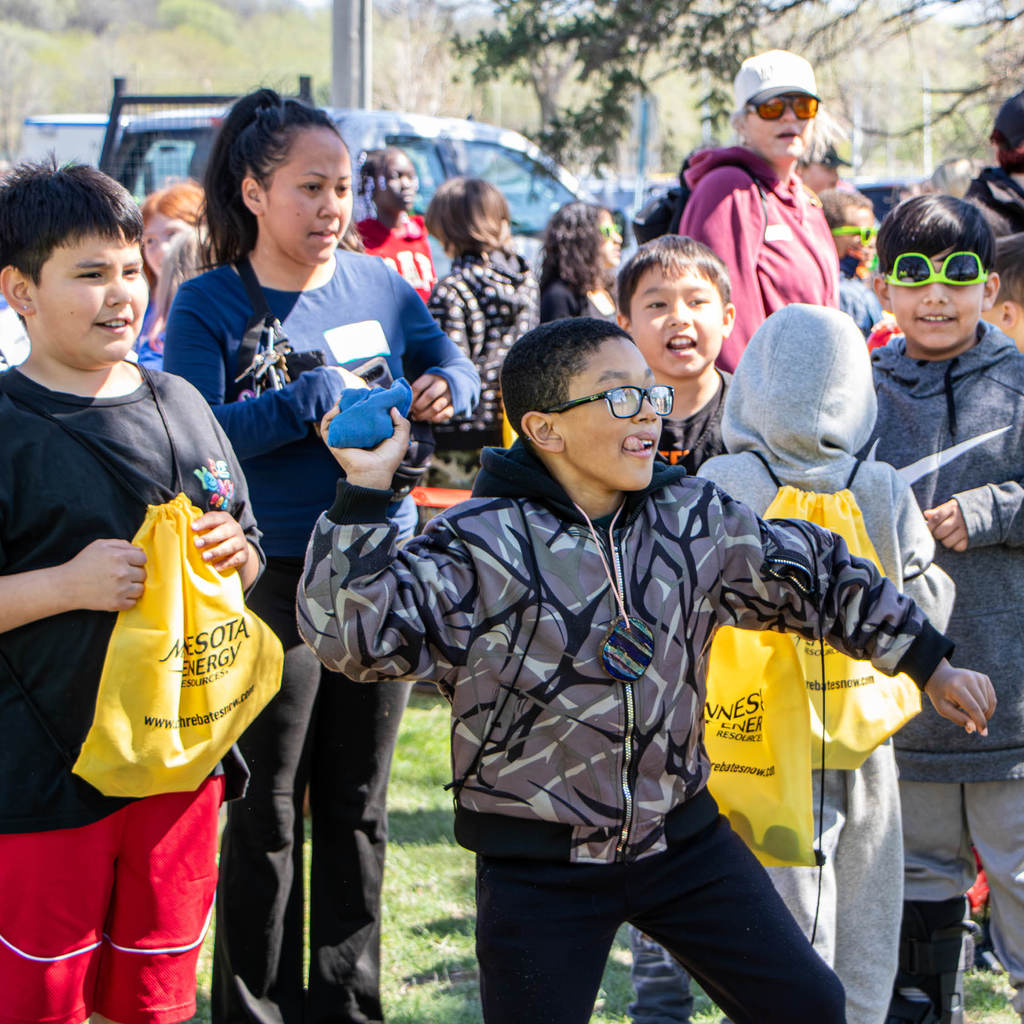 In a crowd, a boy reels back to throw a bean bag and sticks out his tongue in consentration.