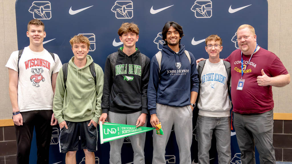 In front of a Century High School backdrop, five male students and a teacher pose for the camera. They are wearing shirts repping the schools they intend to enroll in after high school.