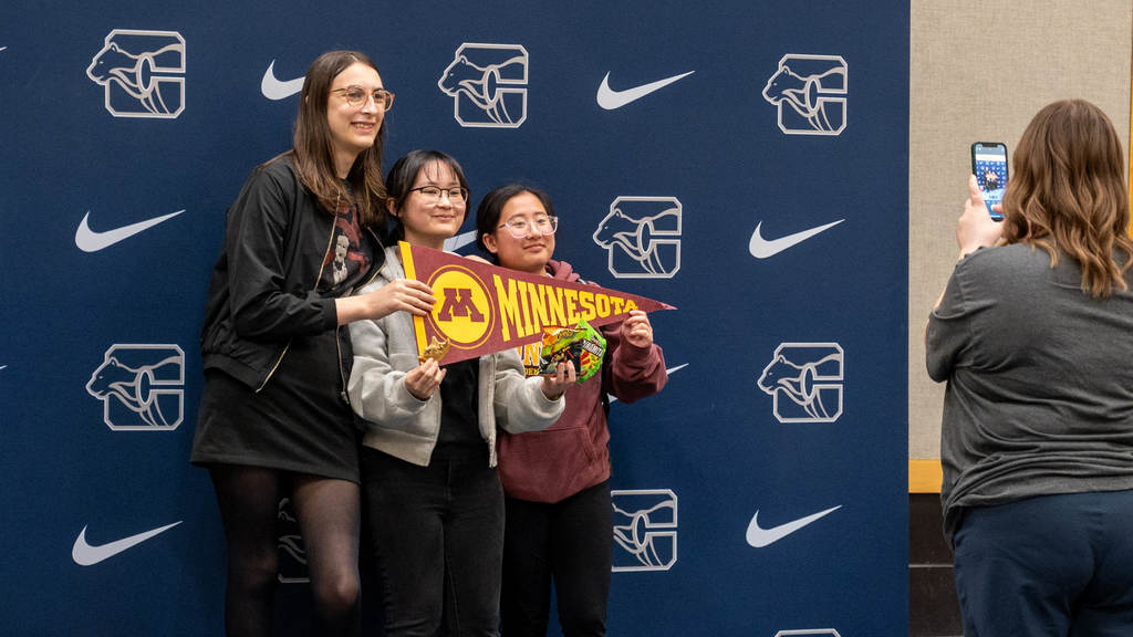 In front of a Century High School backdrop, three seniors pose with a pennant for the U of M. 