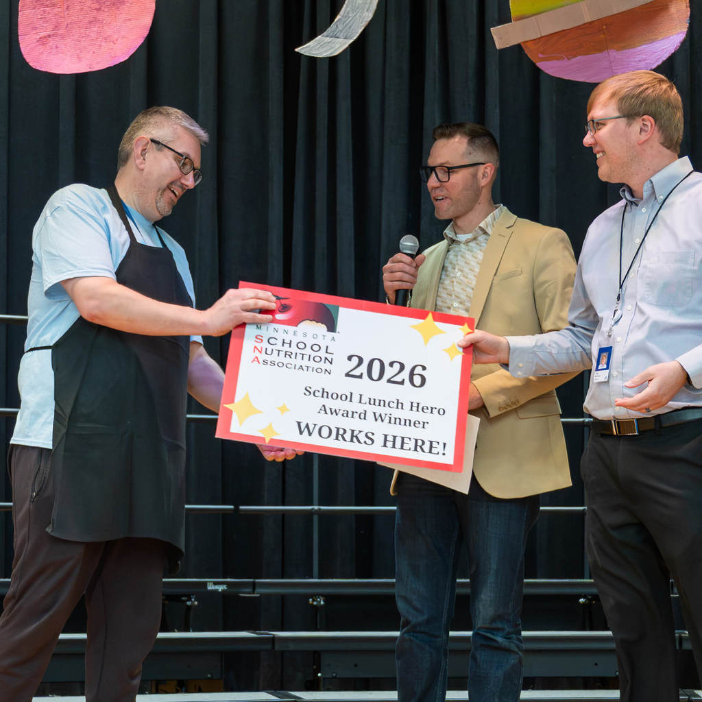 Luke Fenton receives an award (symbolized by a sign) for School Lunch Hero Award by the Minnesota Student Nutrition Association from the Director of Student Nutrition at Rochester Public Schools, Dale Winham.