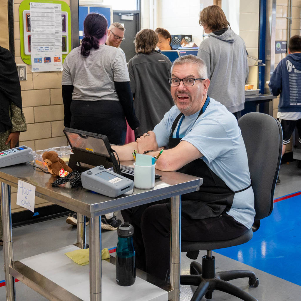 A Student Nutrition Services Associate sits at the payment station with a tablet, pencil holder, and owl plush. Behind him are students in the lunch line.