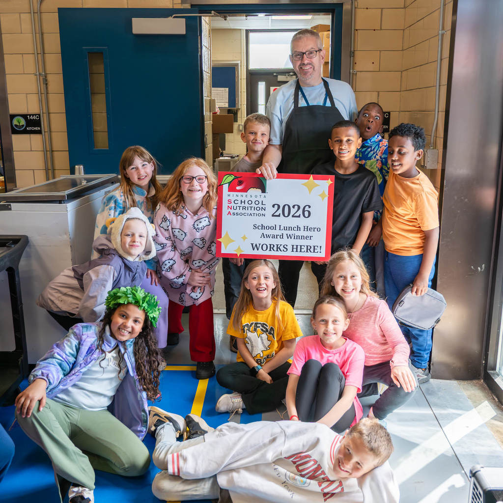 In a cafeteria, an employee stands holding a sign that says, "Minnesota School Nutrition Association 2026 School Lunch Hero Award Winner works here!" He is surrounded  by adoring kids and they all smile at the camera.