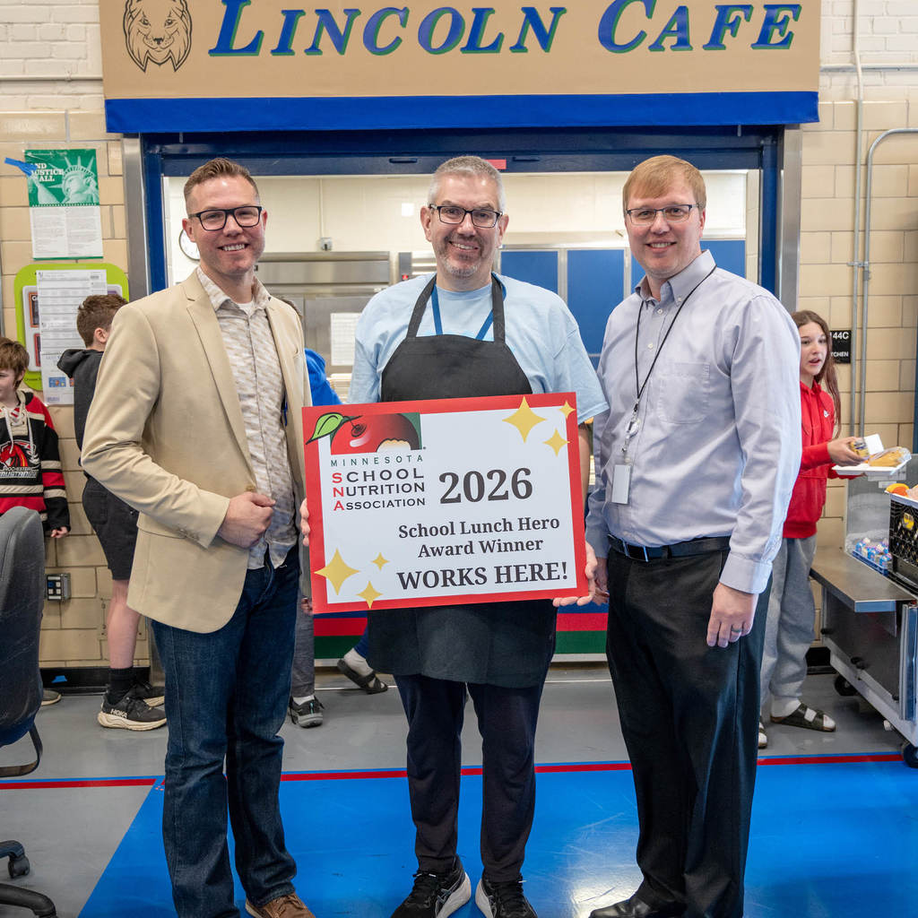 Brent from the Minnesota Student Nutrition Association, Student Nutrition Associate, Luke Fenton, and Director of Student Nutrition at Rochester Public Schools, Dale Winham stand in the cafeteria. Behind them a sign reads, "Lincoln Cafe." Luke holds a sign reading, "Minnesota School Nutrition Association 2026 School Lunch Hero Award Winner works here!" 
