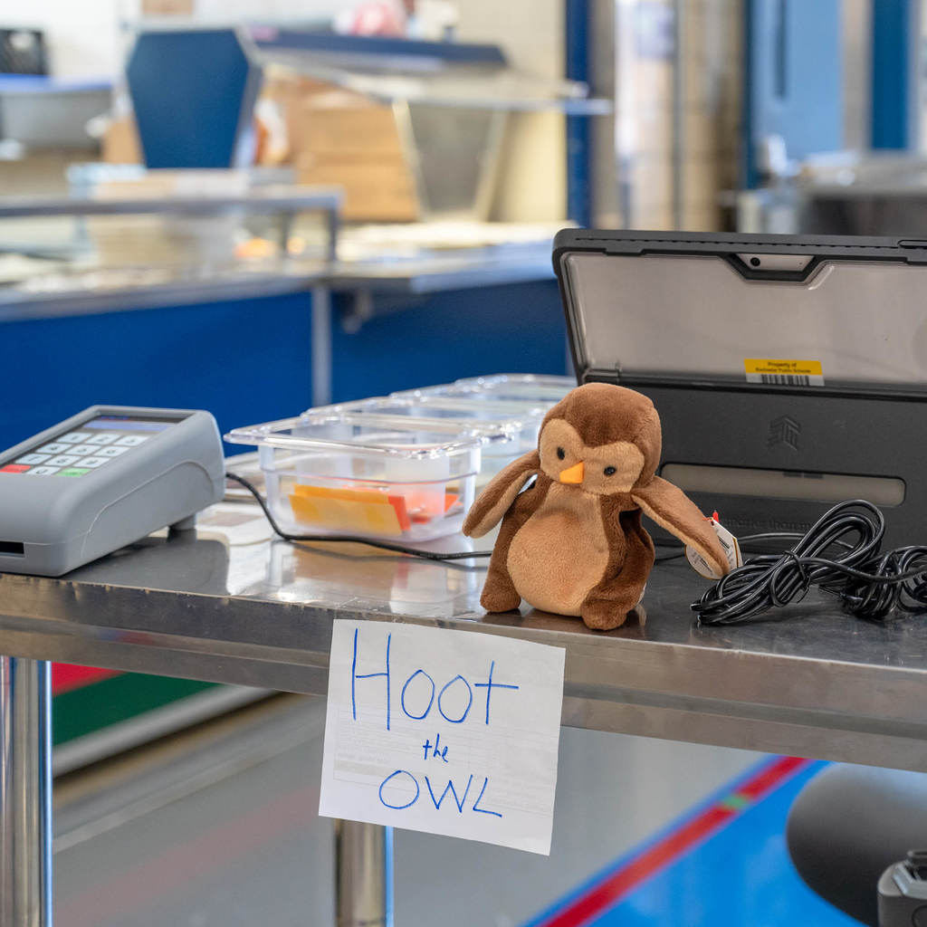 A close up view of the payment station in front of a school kitchen. On the station there are bins, a tablet, codes, a plush brown owl, and a sign that reads, "Hoot the Owl."