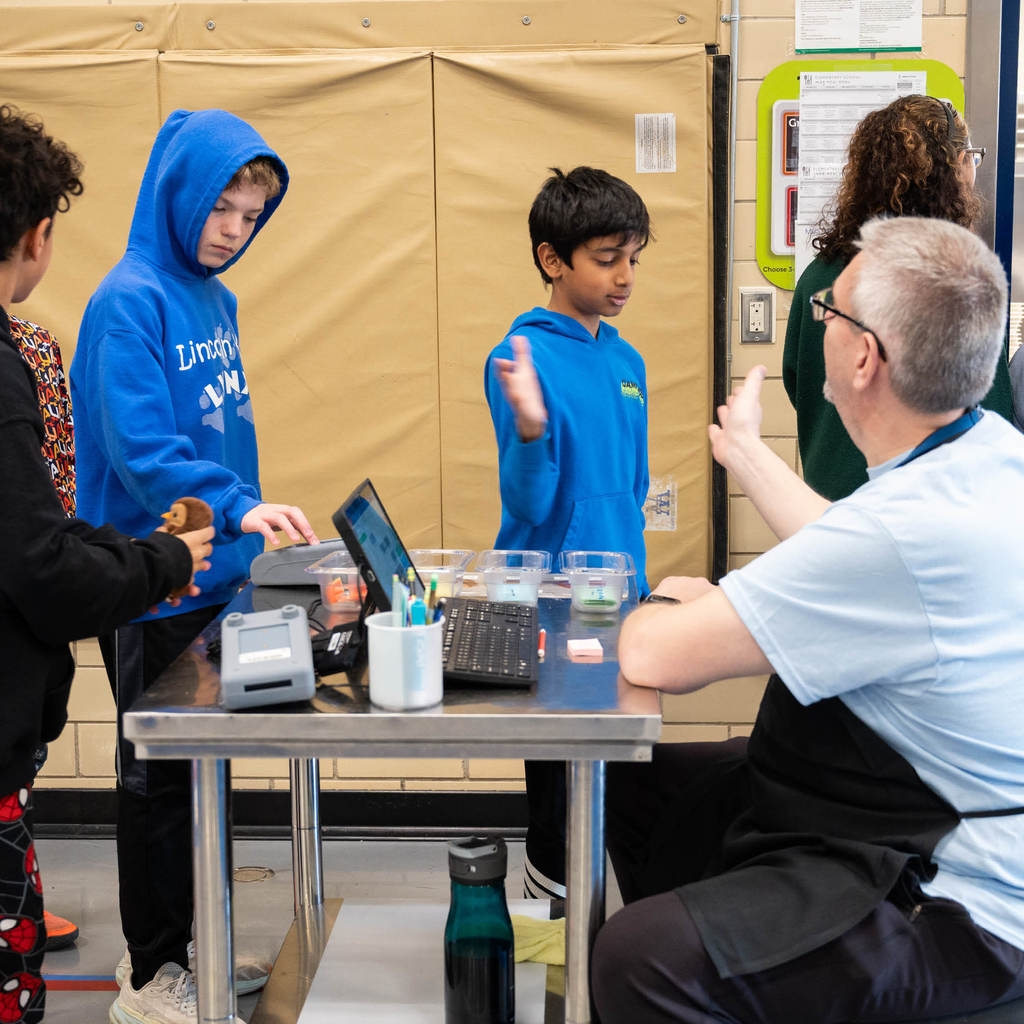 In a lunchline, a boy in a black hoodie holds a plush owl, a boy in a blue hoodie types in his student code at the pay station, and another boy high-fives a Student Nutrition Services Associate.