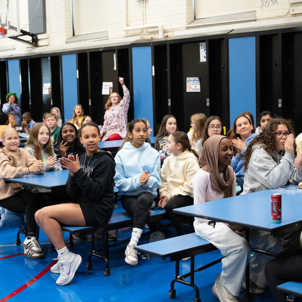 In a cafeteria, middle school students sit at blue tables and clap for the award-winner on the stage (not in frame).