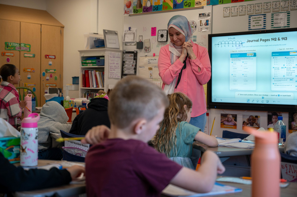 Teacher wearing pink and a teal hijab at the front of the classroom smiling down at her students as they work through a problem in their journals.