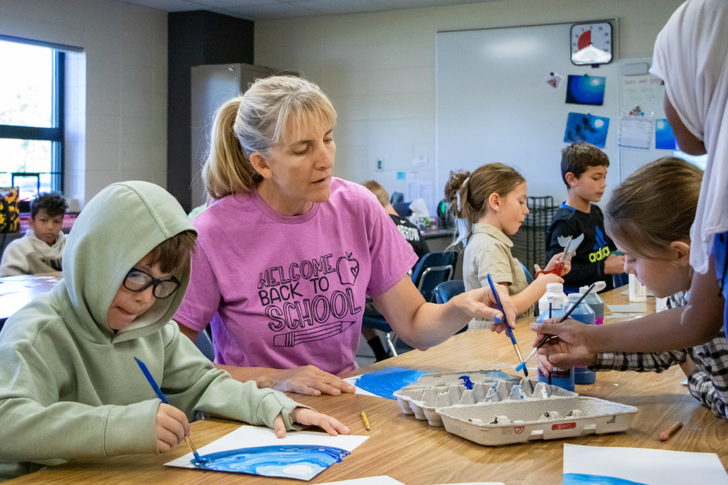 Older female art teacher with grey hair painting alongside a student as they work on their water project.