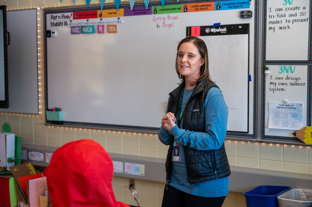 Female teacher in front of the classroom smiling as she listens to a question from a student.